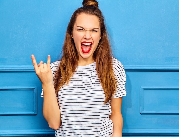 Modèle de jeune femme élégante dans des vêtements d'été décontractés avec des lèvres rouges, posant près du mur bleu. Crier et montrer le signe du rock and roll