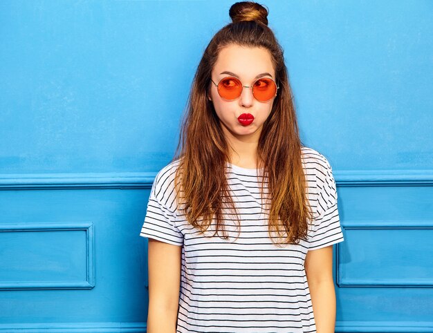 Modèle de jeune femme élégante dans des vêtements d'été décontractés avec des lèvres rouges, posant près du mur bleu. Souffler ses joues
