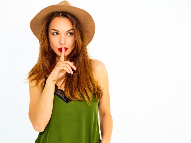 Modèle jeune femme élégante dans des vêtements d'été vert décontracté et chapeau brun avec des lèvres rouges montrant le geste de silence avec le doigt, isolé