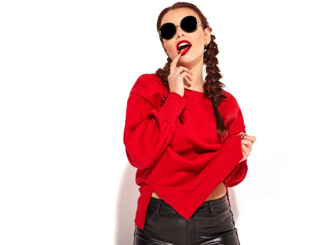 modèle jeune femme souriante heureuse avec maquillage lumineux et lèvres colorées avec deux nattes et lunettes de soleil dans des vêtements d'été rouges isolés. toucher sa bouche