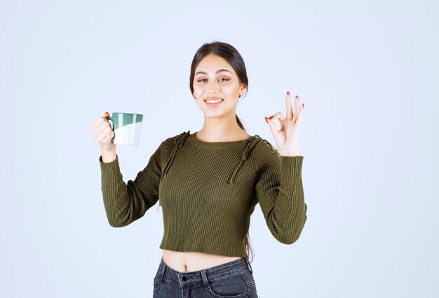 Un modèle de jeune femme souriante tenant une tasse et montrant un geste correct.