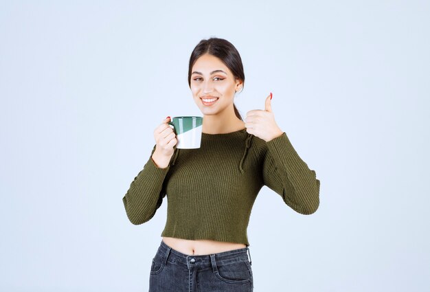 Un modèle de jeune femme souriante tenant une tasse et montrant un pouce vers le haut.