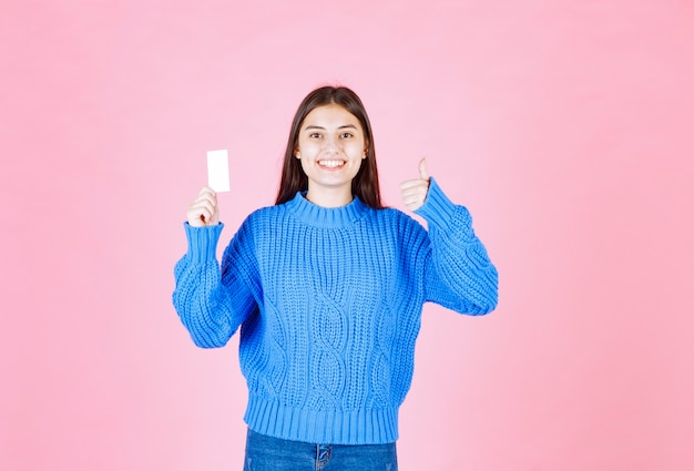 modèle de jeune fille souriante avec une carte montrant un pouce vers le haut sur un mur rose.