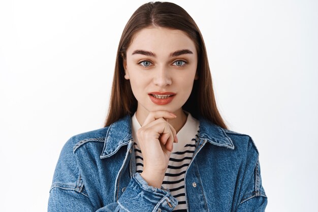 Le modèle de jeune fille touche le menton, a l'air déterminé et réfléchi à l'avant, pense à quelque chose, visage pensif, se concentre tout en faisant un choix, debout sur un mur blanc