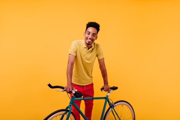 Modèle masculin africain intéressé en pantalon rouge souriant. Portrait de mec noir inspiré debout près de vélo vert.