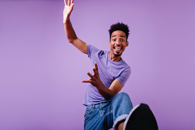 Modèle masculin drôle bouclé sautant et riant. Joyeux jeune homme en t-shirt et jeans dansant.