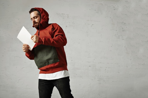 Un modèle masculin en jeans, t-shirt blanc et parka rouge et gris lisant quelque chose sur une feuille de papier vierge blanc isolé sur blanc