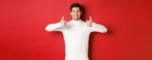 Modèle masculin joyeux et beau en pull blanc, montrant le pouce levé en signe d'approbation, comme quelque chose de bien, debout sur fond rouge et souriant heureux.