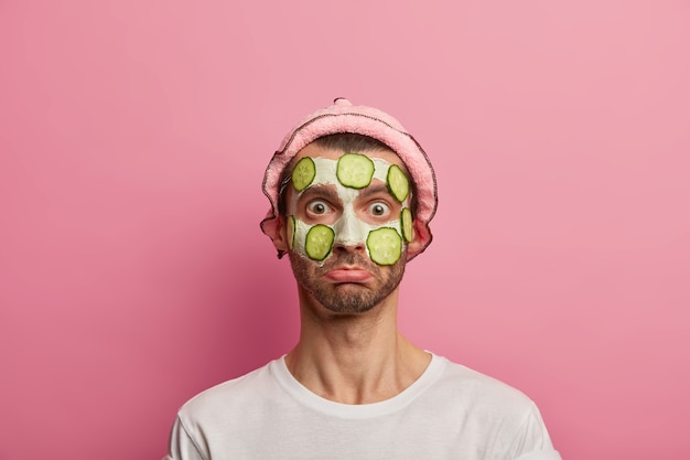 Un modèle masculin perplexe applique un masque facial à l'argile et des tranches de concombre pour rajeunir la peau, porte un chapeau de bain, un t-shirt blanc