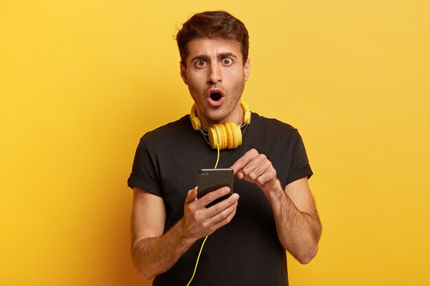 Un modèle masculin stupéfait montre l'écran du téléphone intelligent, lit une notification étrange, une chanson perplexe a disparu de la liste de lecture