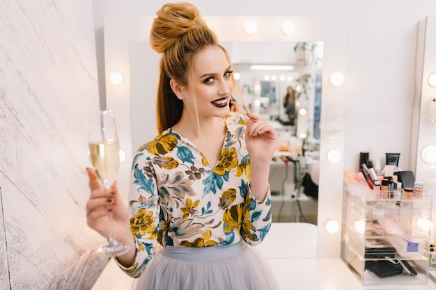 Modèle à la mode attrayant avec coiffure de luxe, beau maquillage souriant à la caméra dans un salon de coiffure avec une coupe de champagne