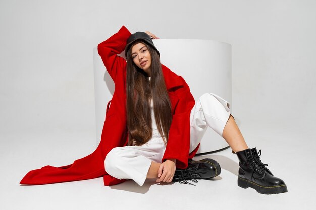 Modèle à la mode en chapeau élégant, manteau rouge et bottes posant sur un mur blanc en studio