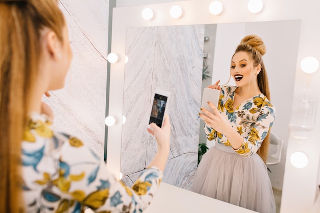 Modèle à la mode avec coiffure élégante, maquillage professionnel faisant selfie dans un miroir dans un salon de coiffure