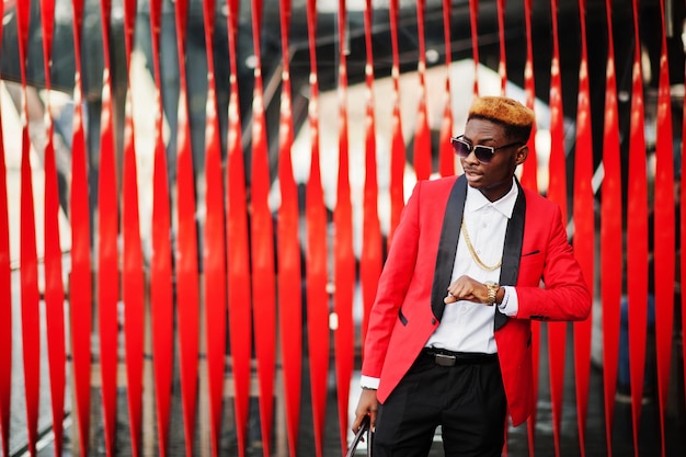 Modèle de mode homme afro-américain en costume rouge avec cheveux et sac à main en surbrillance posés dans la rue et regardant ses montres