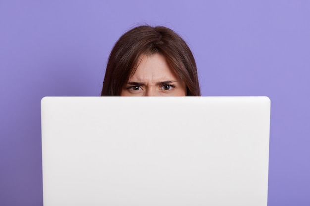 Modèle se cachant derrière un ordinateur portable isolé sur un mur lilas, ayant une expression faciale en colère, femme aux cheveux noirs derrière un carnet de notes blanc, pigiste pendant le travail.