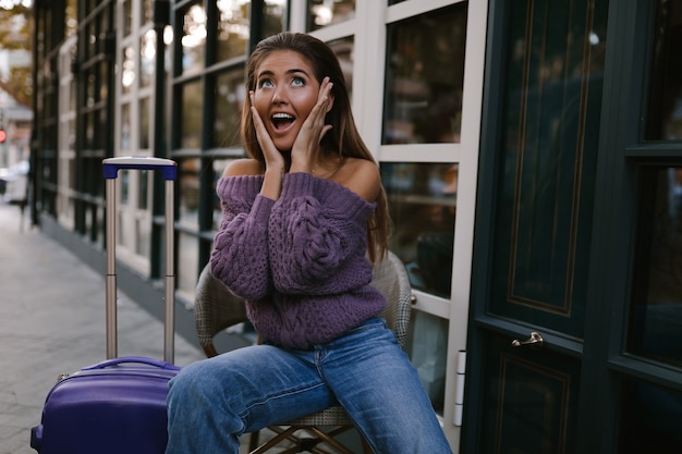 Modèle sorti avec la valise bleue assise sur la chaise près du café, dans le pull violet, jeans, maquillage, coiffure, émotions, surpris, automne, blonde, heureuse, tricotée, souriante