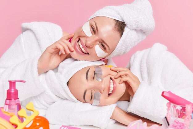 les modèles féminins sourient avec joie les têtes inclinent appliquent des patchs de beauté sous les yeux aime les procédures de soins de la peau vêtus de peignoirs blancs et doux isolés sur rose