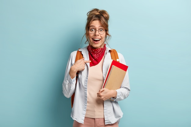 Moi, vraiment? Heureuse femme européenne souriante se montre, sourit largement, ne peut pas croire à un examen réussi, pose avec le bloc-notes