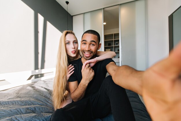 Moments drôles de joli couple s'amusant, faisant selfie sur le lit à la maison dans un appartement moderne. Femme aux longs cheveux blonds, vraies émotions, amoureuse, femme, mari, relation