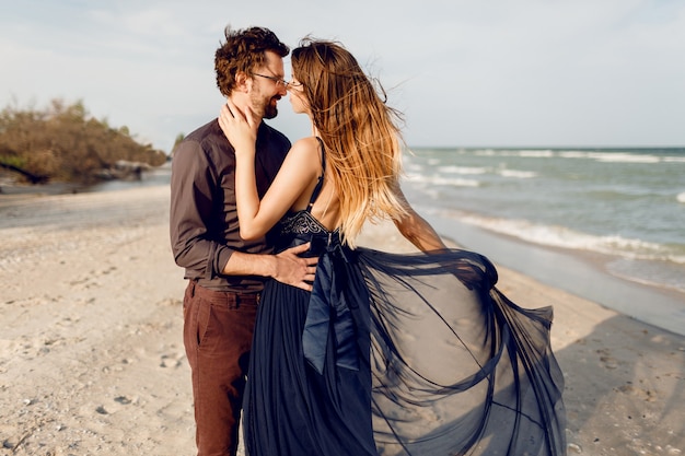 Moments romantiques de beau couple, femme à la mode et homme posant en plein air près de la mer. Robe bleue incroyable et tenue décontractée. Vacances de noces.