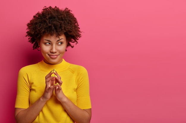 Photo gratuite mon plan est parfait. jolie femme afro-américaine planifie quelque chose, steepls doigts et regarde avec une expression rusée de côté, sourit sournoisement, pose sur un mur rose, copiez l'espace de côté pour votre promo