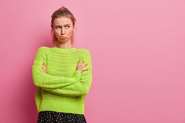 Moody femme offensée se tient avec les mains croisées, porte les lèvres, être offensé, regarde sous le front, porte un pull vert, se dresse sur un mur rose