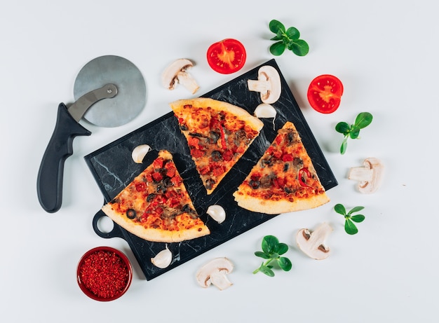 Morceaux de pizza à la tomate et à l'ail, épices, champignons, feuilles de menthe et un coupe-pizza dans une planche à découper sur fond bleu clair, à plat.