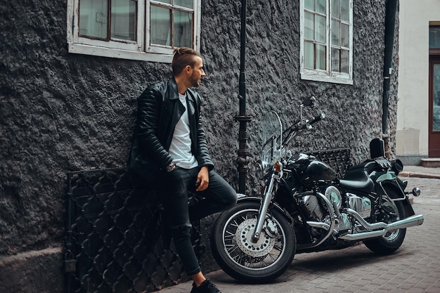 Motard à la mode vêtu d'une veste en cuir noir et d'un jean appuyé sur un mur près de sa moto rétro dans une rue de la vieille Europe.