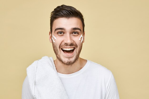 Émotionnel excité jeune homme barbu posant isolé en t-shirt et serviette sur son épaule va se raser et prendre une douche après l'entraînement en salle de gym, rayures de mousse blanche sur ses joues