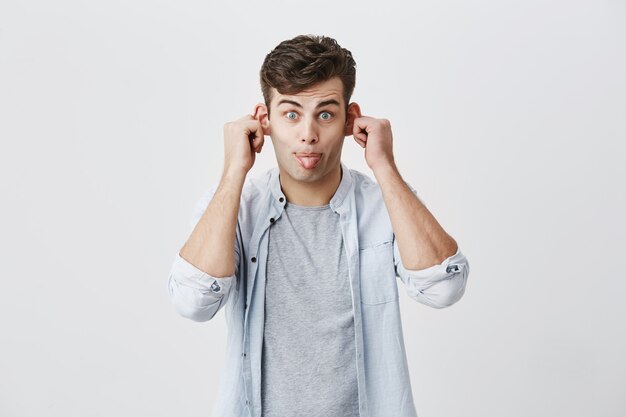 Émotionnel jeune étudiant masculin en chemise bleue sur un t-shirt gary grimaçant, se moquant, faisant des grimaces, tirant ses oreilles, sortant sa langue, regardant avec une expression stupide sur son visage.