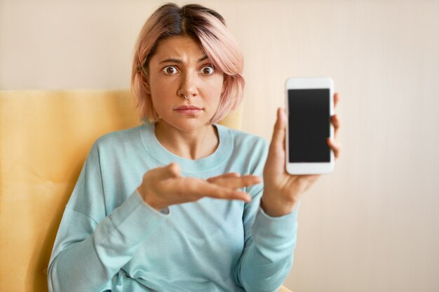 Émotionnelle jeune femme aux cheveux rose regardant la caméra avec choc et surprise, tenant un téléphone mobile