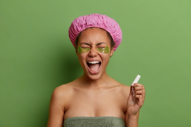 Émotionnelle jeune femme crie fort retient le tampon applique un patch de collagène pour réduire les rides enveloppé dans une serviette de bain chapeau imperméable stangs contre le mur vert vif