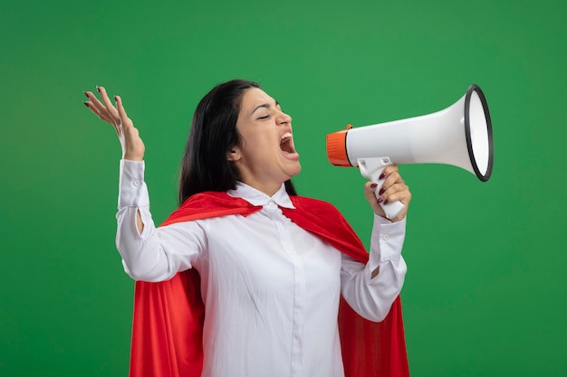 Émotionnelle jeune superwoman debout en vue de profil en criant dans le haut-parleur à tout droit isolé sur mur vert