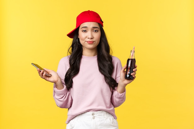 Émotions des gens, boissons et concept de loisirs d'été. Jolie fille asiatique indécise et désemparée au bonnet rouge, tenant une bouteille avec du soda et un téléphone portable, haussant les épaules, incertain, fond jaune.