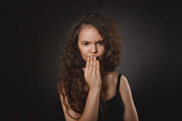 Émotions humaines négatives et expressions faciales. Photo de frustré séduisante jeune femme aux cheveux noirs en haut noir fronçant les sourcils et tenant la main sur ses lèvres, souffrant de maux de dents