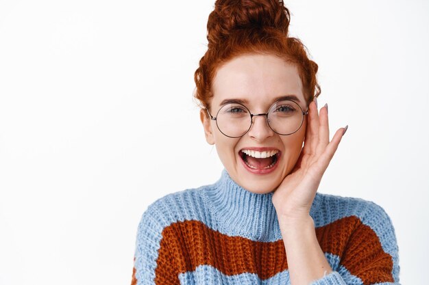 Émotions positives. Heureuse rousse dans des verres en riant sans soucis. Femme aux cheveux roux et lunettes touchant son visage, souriant sur blanc