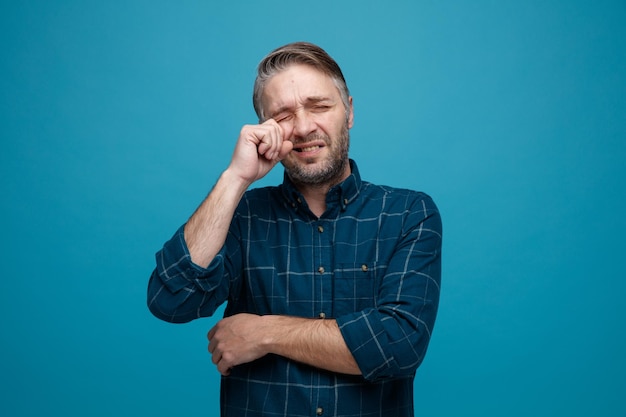 Moyen-âge triste homme contrarié aux cheveux gris en chemise de couleur foncée pleurant en se frottant les yeux avec la main debout sur fond bleu