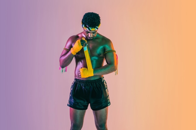 Photo gratuite muay thaï. jeune homme exerçant la boxe thaï sur un mur dégradé à la lumière du néon. combattant pratiquant, entraînement aux arts martiaux en action, mouvement. mode de vie sain, sport, concept de culture asiatique.