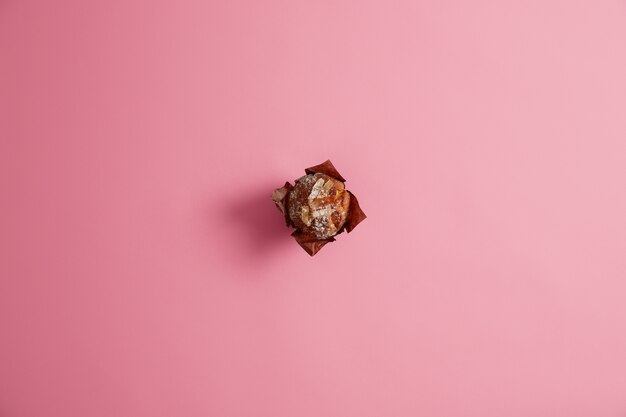 Muffin cuit prêt en poudre avec du sucre dans du papier brun isolé sur fond rose. Confiserie fraîche, douce vie, concept de malbouffe. Petit déjeuner du matin. Dessert pour les gourmets. Mise au point sélective.