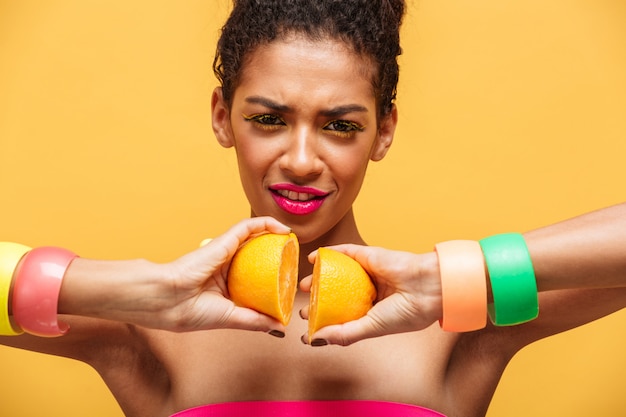 Mulâtre élégant conceptuel femme avec du maquillage multicolore reconnectant deux parties d'orange fraîche dos ensemble isolé, sur mur jaune