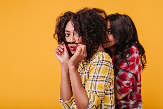 Mulâtre joue avec les cheveux, faisant de la moustache avec une mèche, tandis que son amie se cache derrière son dos. Portrait de filles joyeuses s'amusant.