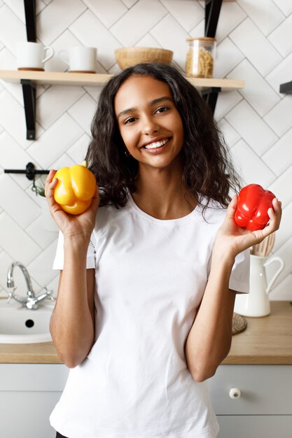 Mulâtre souriante vêtue d'un t-shirt blanc, avec un joli visage et des cheveux lâches, tient des poivrons jaunes et rouges dans les mains dans la cuisine