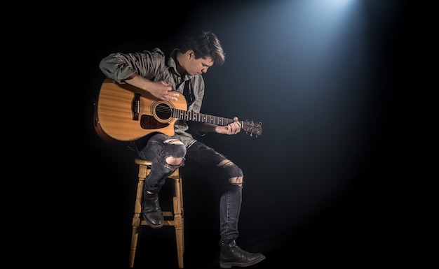 musicien jouant de la guitare acoustique, assis sur une chaise haute, fond noir avec une belle lumière douce