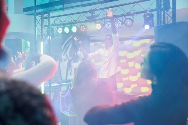 Photo gratuite musicien avec microphone levant la main tout en divertissant la foule de la scène en boîte de nuit. dj chantant et jouant à un spectacle de musique électronique et à une soirée discothèque dans un club avec des projecteurs