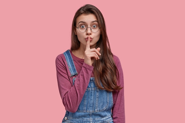 Mystérieuse belle jeune femme fait un geste chut, porte une salopette en denim, demande de ne pas dire son secret, a des lunettes optiques