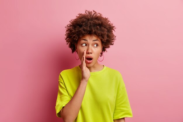Une mystérieuse femme aux cheveux bouclés choquée garde la main près de la bouche ouverte dit que le secret se propage des rumeurs murmure quelque chose de secrètement concentré de côté porte un t-shirt décontracté isolé sur un mur rose