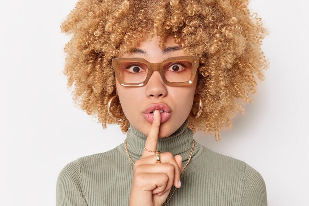 Mystérieuse jeune femme aux cheveux bouclés appuie l'index sur les lèvres fait un geste de silence demande à être calme porte des lunettes et un col roulé isolé sur fond blanc. Chut tais toi s'il te plait