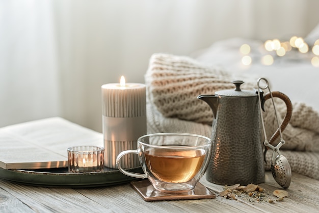 Nature morte confortable avec une tasse de thé en verre, une théière et des bougies sur un arrière-plan flou avec bokeh.