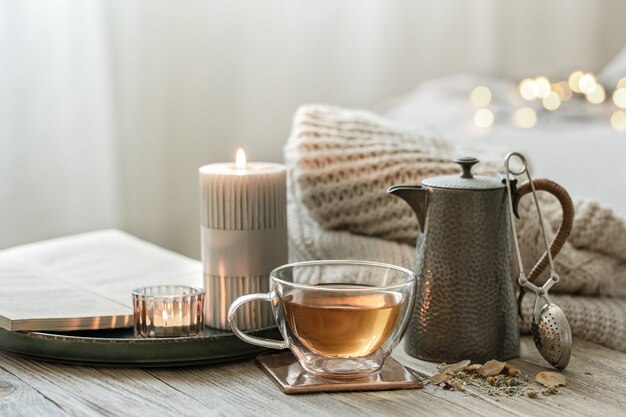 Nature morte confortable avec une tasse de thé en verre, une théière et des bougies sur un arrière-plan flou avec bokeh.