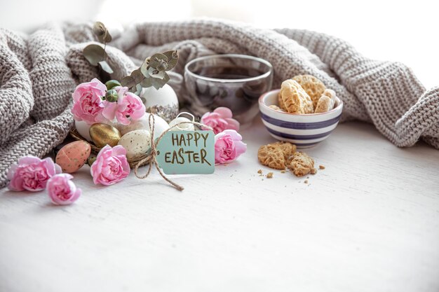 Nature morte avec du thé, des biscuits, des œufs, des fleurs et l'inscription Joyeuses Pâques sur la carte postale